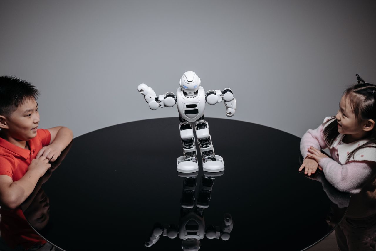 hero-img-01 Two children interacting with a robot toy on a table indoors, showcasing technology and fun.